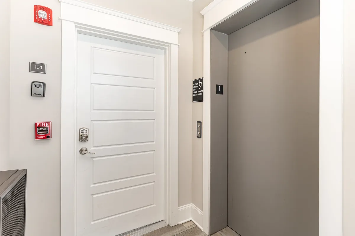 The photo features the main entrance door, a modern elevator, fire alarm, and a room number sign. The space is clean and welcoming, conveying the first impression guests have when they enter the property and make use of the elevator to access their units.