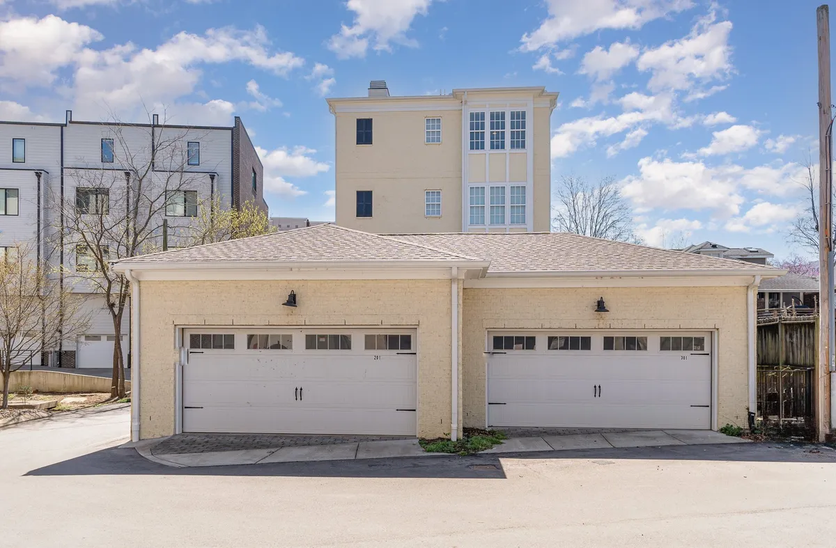 This image shows the exterior of the building with two garage doors and upper-level windows, surrounded by trees. The driveway area in front of the garages is visible, suggesting ample parking space for multiple vehicles.
