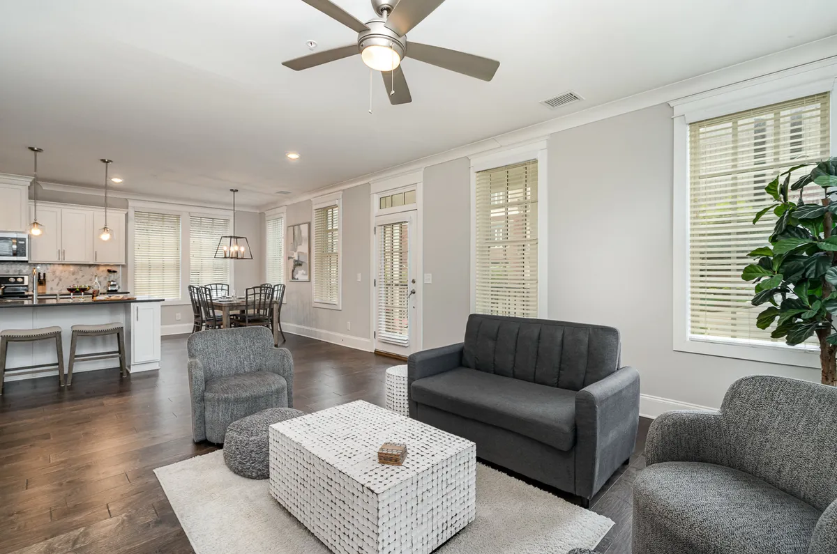 A comfortable and modern living room space with a plush sofa, coffee table, and large windows drawing in sunlight. The kitchen area is visible in the background, underscoring the open layout ideal for socializing and family time.