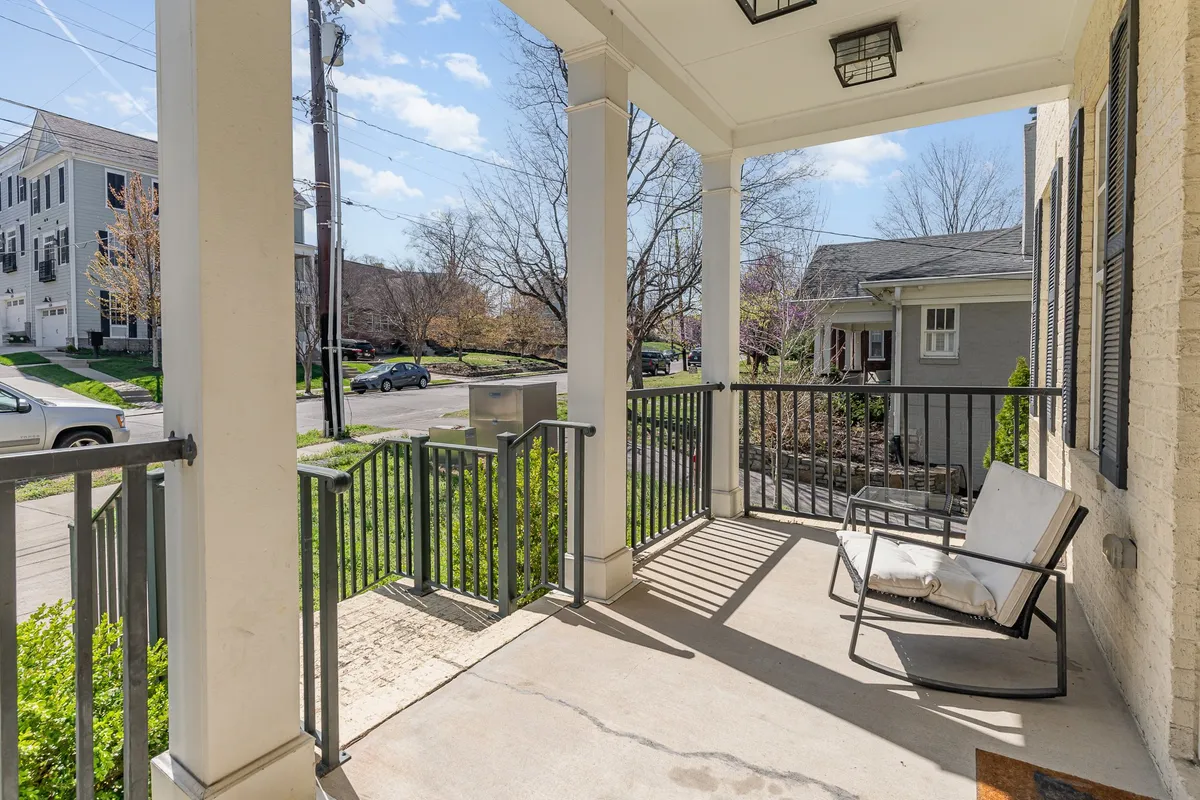 A classic front porch scene featuring a welcoming porch swing, elegant decorative columns, and lush greenery framing a view of the quiet street. The porch is shaded by mature neighborhood trees, creating an inviting spot to enjoy the outdoors.