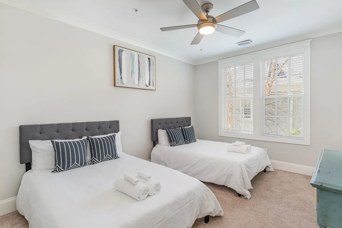 A guest bedroom with two stylish beds featuring gray headboards and decorative pillows, accompanied by modern artwork and a window with blinds. The room is bright and flexible, ideal for families or friends traveling together.