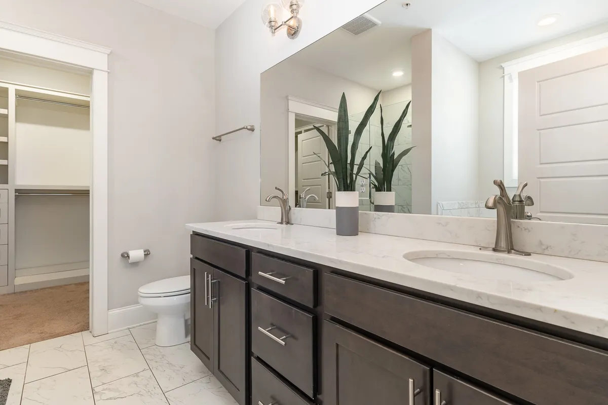 A spacious and modern bathroom with a double sink vanity, large mirror, houseplants, and an adjoining walk-in closet. The tranquil design and thoughtful amenities evoke a spa-like experience.